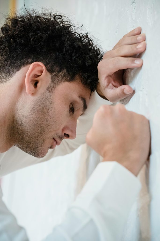 Side profile of a contemplative man with curly hair and clenched fists leaning against a white wall.